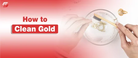 Hand using a soft toothbrush to clean gold jewelry in a small bowl of warm soapy water.